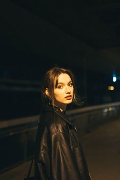 a woman standing in the dark with her hand on her hip wearing a black leather jacket