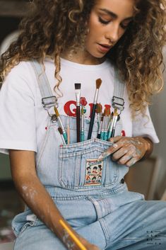 a woman sitting on the floor with lots of paint brushes in her pocket and holding a pencil