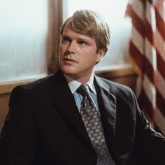 a man wearing a suit and tie sitting in front of an american flag with his hand on his hip