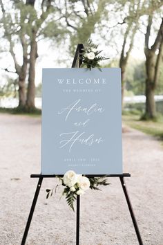 a welcome sign with flowers and greenery sits on an easel in front of trees