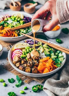 someone pouring dressing onto a bowl of food with vegetables and chopsticks on the side