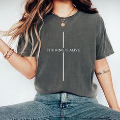 "Discover serenity and inspiration with our \"The King is Alive\" T-Shirt, a wearable testament to faith and hope. This steel blue tee, adorned with a striking white cross formed by the uplifting proclamation \"THE KING IS ALIVE\" and a minimalist vertical line, serves as a subtle yet powerful expression of Christian belief. Crafted for comfort and designed for durability, it's an ideal piece for those who carry their faith close to their heart. Perfect for personal reflection or as a thoughtful Christian Tee Shirts Elevated Faith, Cross Tshirt Designs, Christian Tshirts Designs Woman, Religious Tshirts, Faith Over Fear Shirt, Minimal Shirt Design, Christian Shirts For Women, Faith Tshirts, Personal Reflection