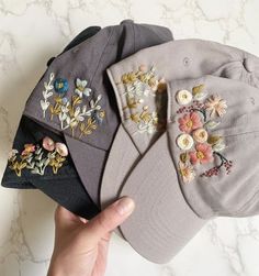 three hats with embroidered flowers on them, one being held up by a person's hand