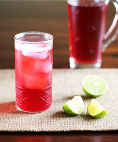 two glasses filled with red liquid next to sliced limes on a cloth covered table