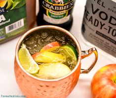 a copper mug filled with liquid and vegetables next to two bottles of vodka, an apple and some other ingredients