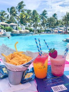 two drinks and some food on a table near a pool with palm trees in the background