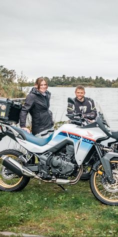 two people standing next to a parked motorcycle near the water on a grassy field with trees in the background