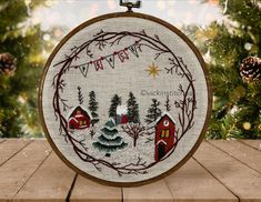 a cross stitch christmas ornament hanging on a wooden table with pine cones and evergreens