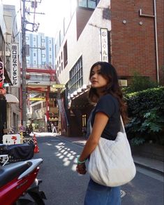 a woman is walking down the street with a bag on her shoulder and a scooter behind her