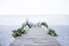 an outdoor ceremony setup with candles and greenery on the sand at the beach in front of the ocean