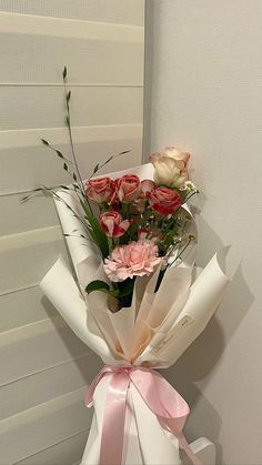 a bouquet of roses wrapped in white paper with pink ribbon on the side, sitting against a wall