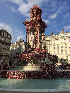 a fountain with flowers all over it and buildings in the backgroung area