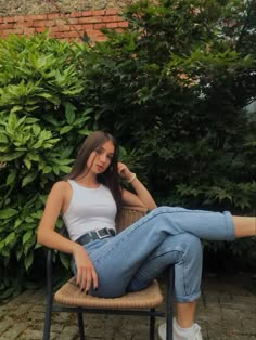 a woman sitting on top of a chair in front of some bushes and trees with her legs crossed