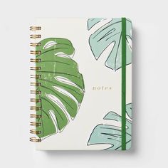 a green and white notebook with palm leaves on the cover, next to a pen