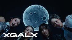 four women are standing in front of a large object with the word x galx on it