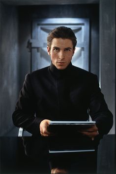 a man in a black suit holding a tablet and looking at the camera while standing in front of a door