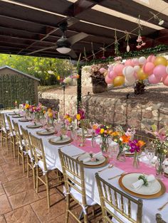 a long table set up for a party with balloons and flowers on the wall behind it