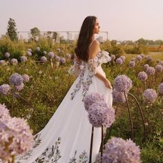 a woman is standing in a field with purple flowers and wearing a white dress that has floral embroidery on it