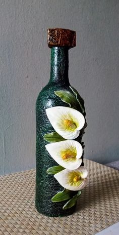 a green vase with three white flowers in it sitting on a woven table cloth next to a gray wall