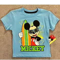 This Fun Disney Mickey Mouse T-Shirt Is Perfect For Little Beach Lovers! Available In Many Sizes, It Features A Cute Surfing Beach Theme That Any Toddler Boy Will Love. Made With Quality Materials And Featuring The Beloved Disney Character, This T-Shirt Is Sure To Become A Favorite. Playful Cartoon Print T-shirt For Disney Trips, Disney Summer Cartoon Print T-shirt, Disney Character Print Summer T-shirt, Themed Short Sleeve Tops For Disney Trips, Disney Crew Neck T-shirt For Summer, Blue Character Print T-shirt For Summer, Casual Mickey Mouse Tops For Playtime, Cotton Mickey Mouse Top For Playtime, Playful Mickey Mouse Tops For Playtime