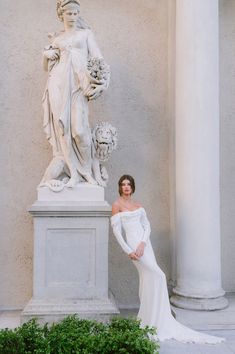 a woman standing next to a statue in front of a white building with columns and greenery