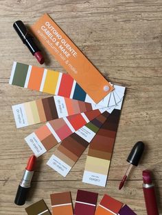 several shades of paint and lipstick on a wooden table with an orange card next to them