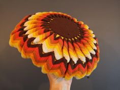 a hand holding a knitted sunflower in the shape of a flower with brown, yellow and orange petals