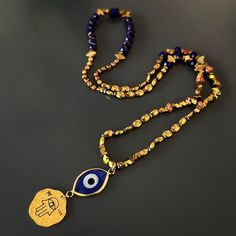 Good Luck Evil Eye and Hamsa Necklace, is a stunning fusion of cultural symbolism and elegant design. This captivating necklace is meticulously crafted, featuring a combination of gold-colored hematite stone beads, blue crystal beads, a sterling silver Hamsa pendant plated with 18k gold, and an 18k gold-plated evil eye glass pendant. At the heart of the necklace lies a sterling silver Hamsa pendant, plated with 18k gold. The Hamsa symbolizes protection and blessings in many cultures, serving as Spiritual Necklace With Colorful Beads For Festive Occasions, Gold Jewelry With 108 Round Beads, Symbolic Gemstone Bead Necklaces For Gifts, Beaded Amulet Jewelry For Meditation, Meditation Beaded Amulet Jewelry, Hand-strung Beaded Necklaces As Festival Gifts, Bohemian Gold Necklace With Polished Beads, Hand-strung Yellow Gold Beaded Necklace As Gift, Artisan Brass Necklace With Polished Beads