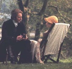 a man and woman sitting on lawn chairs talking to each other in the grass with trees behind them