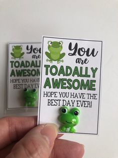 someone is holding two green frog brooches in their left hand and one has the words you are toadally awesome, hope you have the best day ever