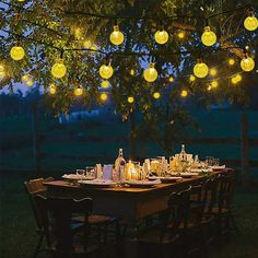 an outdoor dinner table set up with candles and lights strung from the tree over it