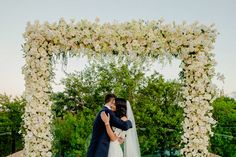 a bride and groom embracing each other in front of an outdoor wedding arch with white flowers