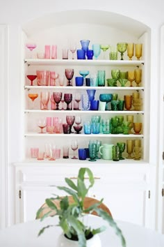 a white shelf filled with lots of colorful glasses on top of a table next to a potted plant