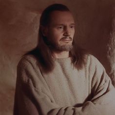 a man with long hair and beard wearing a white sweater standing in front of a stone wall