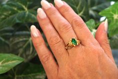 a woman's hand holding a ring with green and yellow stones on it in front of a potted plant