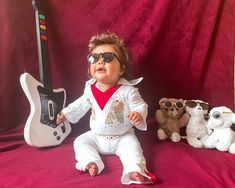 a baby wearing sunglasses and sitting next to stuffed animals with a guitar in the background