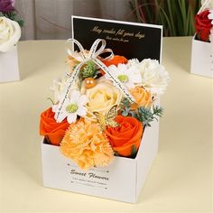 an orange and white flower arrangement in a card box on a table with other flowers