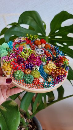 a hand holding a wooden plate with colorful beaded decorations on it and a plant in the background