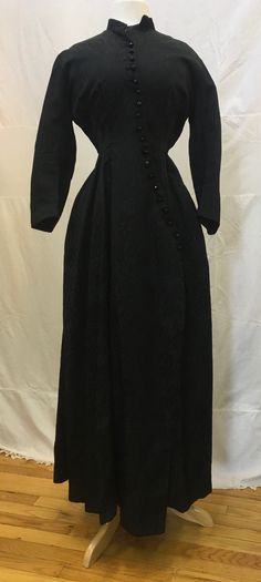 "1880s, 36\" bust, black silk embossed silk full length \"wrapper\", at home dress.  The wrapper has long sleeves and a small high collar.   The wrapper is fastened in front with 19 jet buttons sewn diagonally, in front, from neck to hips.  Bodice is lined in tan colored chintz and has 6 boned stays sewn into bodice front and back.  Skirt of dress is open from knee to hem.  Measurements: bust 36\" Waist 26\" Sleeves 18\" long Arm holes 8\" Shoulder to shoulder  16\" Shoulder to hem  51\" Hips 40 Black Victorian Dress With Long Sleeves, Black Long Sleeve Victorian Dress, Formal Black Dress With Historical Design, Regency Style Long Sleeve Victorian Evening Dress, Black Victorian Dress With Historical Design, Formal Black Victorian Dress With Historical Design, Black Long Sleeve Regency Dress, Black Historical Victorian Wedding Dress, Black Victorian Long Sleeve Dress For Evening