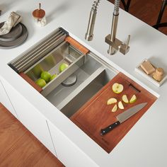 a kitchen sink with cutting board, knife and apples