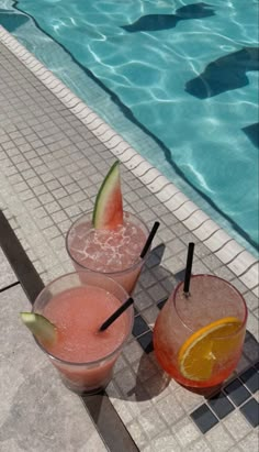 two glasses filled with drinks sitting next to a swimming pool on top of a tiled floor