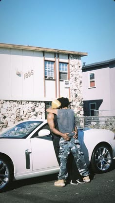 two people standing next to a white sports car in front of a building with a stone wall