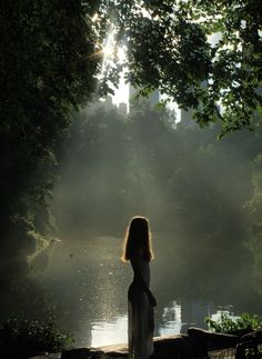 a woman standing in front of a body of water with the sun shining through trees