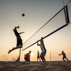 four men playing volleyball on the beach at sunset, with one jumping up to hit the ball