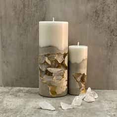 two white candles with gold foil on them sitting next to each other in front of a concrete wall