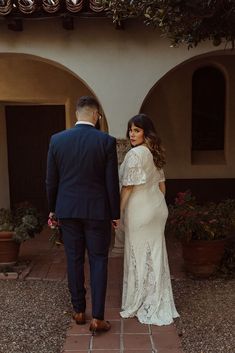 a man and woman standing in front of a building with potted plants on either side