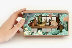 a hand is holding a miniature box with flowers and plants