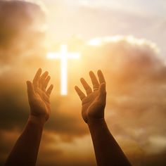 two hands reaching up towards a cross in the sky with sun shining through clouds behind them