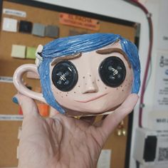 a person holding up a coffee cup shaped like a doll with big eyes and blue hair