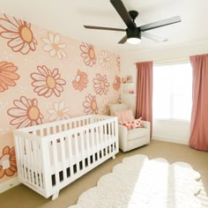 a baby's room with pink and orange flowers on the wall, white crib bedding, and large window